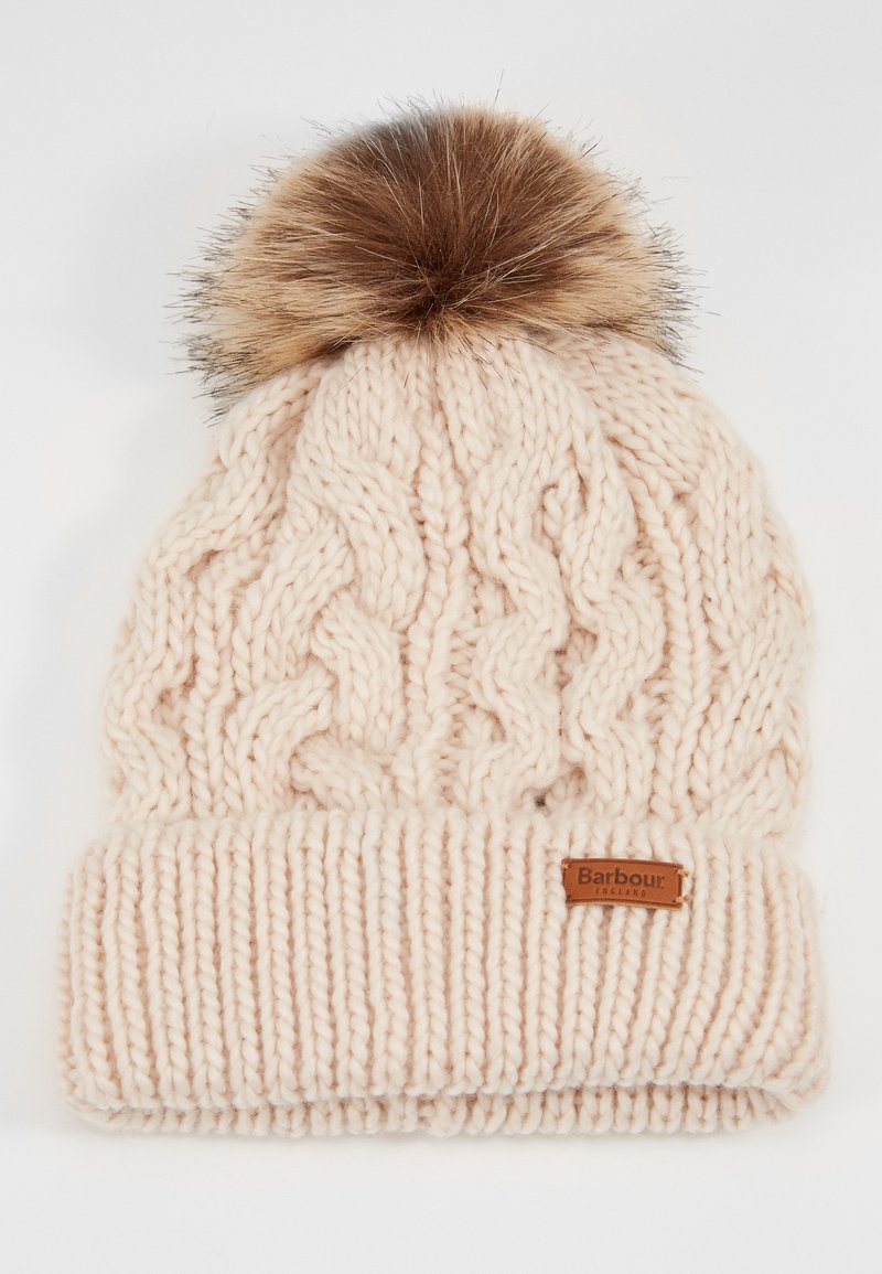 Bonnet d'hiver beige tricoté avec motif torsadé et bord plié, avec un pompon en fausse fourrure marron et une petite étiquette de la marque Barbour.