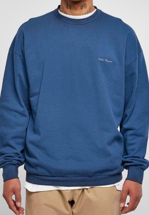 Niebieska bluza oversize z małym białym haftowanym napisem na lewej piersi, noszona na białej koszuli i beżowych spodniach.