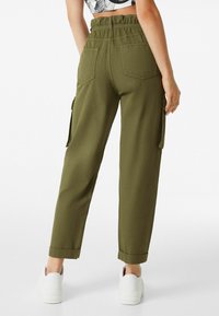 Olivově zelené cargo kalhoty s elastickým páskem, bočními kapsami a ohrnutým lemem, doplněné o bílé tenisky. Hladká textilie.