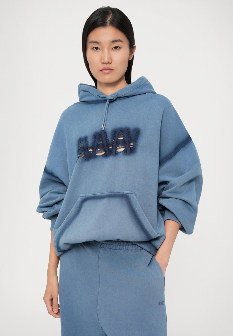 Kvinna med rakt svart hår som bär en lös blå hoodie med texten "WANNA" i struktur och matchande blå träningsbyxor, poserande med handen i fickan.