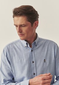 Cămașă button-up de culoare albastră deschisă cu guler, având butoane închise la culoare și un mic logo brodat cu un urs pe partea stângă a pieptului. Textură de material neted.