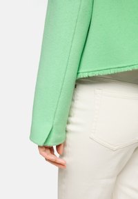 Grüne Fleecejacke mit cropped Design und ausgefranstem Saum. Hand, die auf weißen Jeans ruht und die glatte Textur sowie die schmale Passform zeigt.