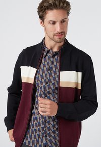 Schwarz-burgunderfarbener Cardigan mit creme- und beigen Streifen. Gerippter Kragen und Bündchen, getragen über einem gemusterten blauen Hemd mit Knopfleiste.