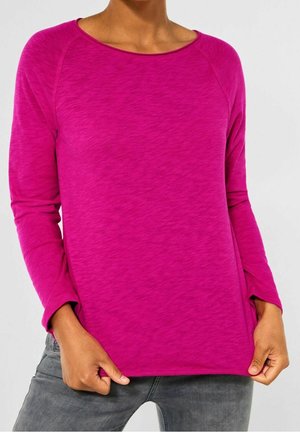 Persoon die een felroze shirt met lange mouwen en grijze spijkerbroek draagt, met handen die de zoom van het shirt iets naar buiten trekken.