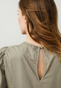 Jasnooliwkowa bluzka z dekoltem w kształcie dziurki, z zapięciem na guzik oraz kontrastowymi szwami na brzegach.
