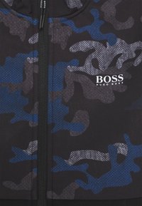 Svart jacka med dragkedja och blå och grå camouflage-mönster. Har en märkeslogotyp "BOSS" på bröstet och strukturerad tyg.