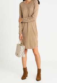 Robe en tricot beige à manches longues avec un col rond, associée à un sac crème texturé avec des accents noirs et des bottines marron.