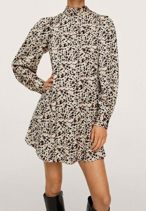 Kobieta ubrana w długą sukienkę z długim rękawem i wysokim kołnierzem w kwiatowy wzór, z bufami na ramionach, oraz czarne kozaki za kolano, stojąca z jedną ręką na biodrze.