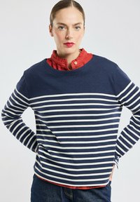 T-shirt à rayures bleu marine et blanches à manches longues avec col rond, superposé sur une chemise rouge à col, associé à un jean en denim bleu.