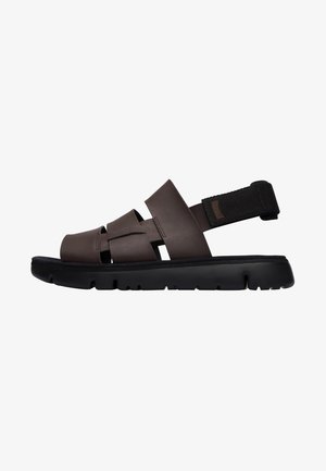 Brown synthetic sandals with a thick black sole, featuring interwoven straps and an adjustable black fabric heel strap for secure fit.