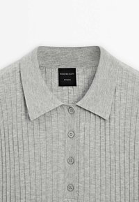 Polo tricoté côtelé gris clair avec col et cinq boutons, montrant une étiquette noire avec texte blanc à l'intérieur du col.