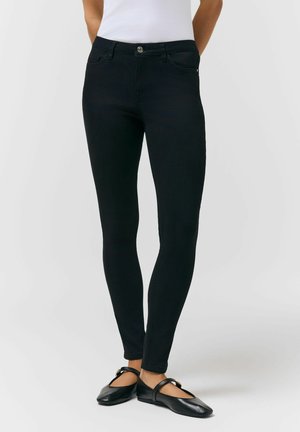 Wąskie czarne spodnie noszone z czarnymi balerinami Mary Jane i białym, bezrękawnym topem, pokazane od talii do stóp na jednolitym tle.