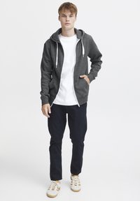 Sudadera gris con cremallera y cordones blancos, llevada sobre una camiseta blanca. Pantalones oscuros y zapatillas claras completan el conjunto, que tiene un ajuste casual.