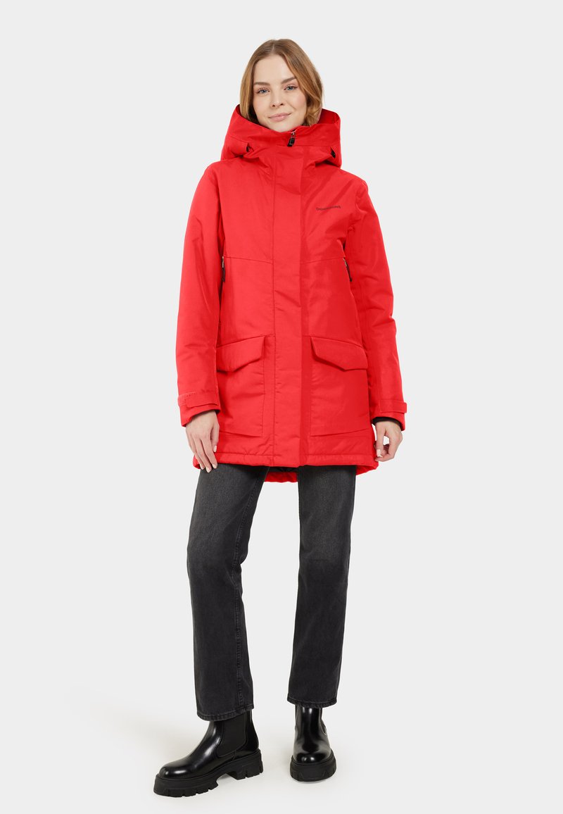 Chaqueta aislante roja con capucha, que presenta bolsillos frontales, cierre con cremallera y elementos ajustables. Combinada con unos vaqueros negros y botas gruesas.