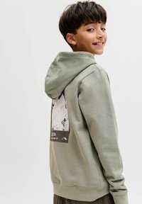 Chlapec sa usmieva cez rameno, má na sebe svetlozelenú mikinu s horskou grafikou na chrbte, pred jednoduchým bielym pozadím.