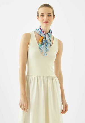 Femme portant une robe crème sans manches et une écharpe colorée à motifs nouée autour du cou, debout devant un fond blanc.