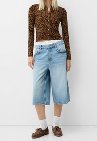 Triko s dlouhým rukávem s leopardím potiskem, hnědá tkanina; modré denimu kalhoty s kapsami, bílý pas, obroubené lemy; hnědé boty na šněrování.