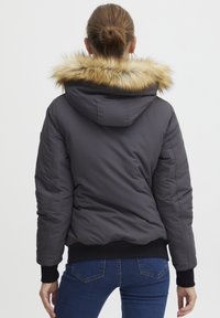 Kort, puffy jakke i mørkegrå med pynt av kunstig pels på hetten. Ribbede mansjetter og kant. Brukt med blå jeans, viser en figursydd silhuett fra baksiden.
