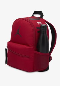 Κόκκινο ύφασμα σακίδιο με μαύρο λογότυπο, φερμουάρ στα διαμερίσματα, και πλαϊνή δίχτυ θήκη που κρατάει μαύρο παγούρι νερού. Περιλαμβάνονται ρυθμιζόμενοι ιμάντες.