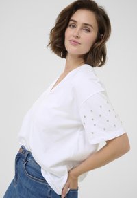 T-shirt blanc à col en V en coton avec des manches courtes, doté d'un motif en perles sur les manches et d'une coupe décontractée et drapée.