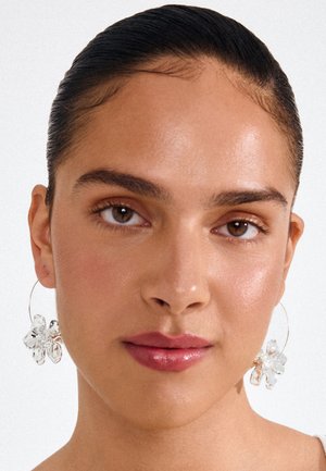 Žena s uhladenými tmavými vlasmi, ktorá nosí priehľadné náušnice v tvare kvetov a svetloružový rúž, pozerá priamo s neutrálnym výrazom.
