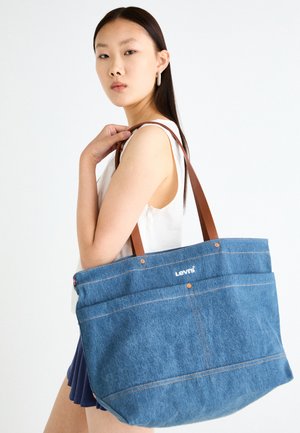 Denim boodschappentas met bruine leren handvatten. Heeft twee grote buitenzakken, witte stiksels en het Levi's logo aan de voorkant.