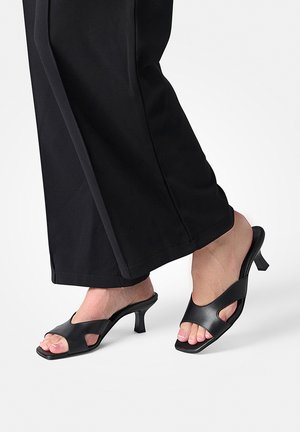 Füße, die schwarze offene Mules mit Absatz und ausgeschnittenen Seiten tragen, kombiniert mit schwarzen Hosen mit weitem Bein auf weißem Hintergrund.