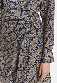 Rochie modelată, de culoare albastru închis, cu un design paisley în nuanțe de aur și alb. Materialul este moale, cu un detaliu legat la talie și mâneci lungi.