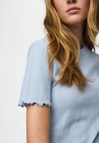 Nainen, jolla on pitkät laineikkaat vaaleanruskeat hiukset, pukeutuneena vaaleansiniseen lyhythihaiseen neulepaitaan, jossa on aaltoilevat reunat hihoissa.