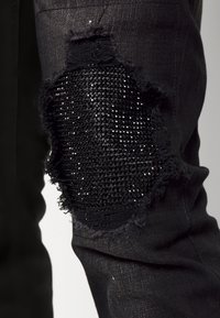 Jeans de mezclilla negros con un gran parche desgastado adornado con rhinestones negros. La tela tiene un ligero brillo, con bordes deshilachados alrededor del agujero.