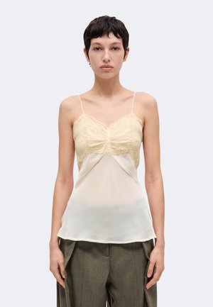 Un mannequin se tient devant un fond clair uni, vêtu d’un caraco en dentelle crème, d’un pantalon court olive et d’escarpins blancs à brides.