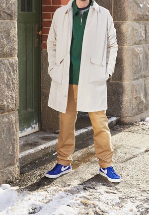 Un homme portant un manteau beige, un pull vert, un pantalon marron clair et des baskets Nike bleues se tient près de l'entrée d'un bâtiment en pierre recouvert de neige.