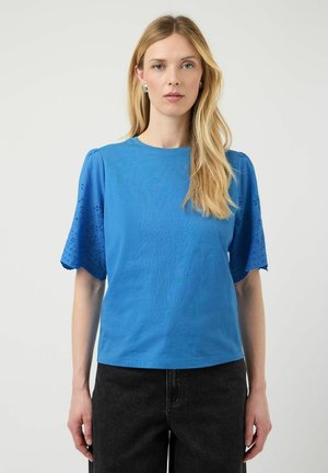 Kvinne med langt blond hår har på seg en blå topp med hullmønstrede pufferm-sleeves og svarte bukser, stående foran en ensfarget lys bakgrunn.