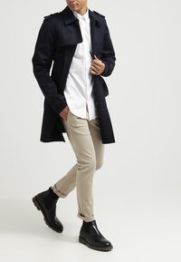 Námořnický trenčkot s ramenními epauletami, bílá košile, béžové chinos kalhoty a černé kotníkové boty s texturovanou podrážkou, stojící v dynamické póze.