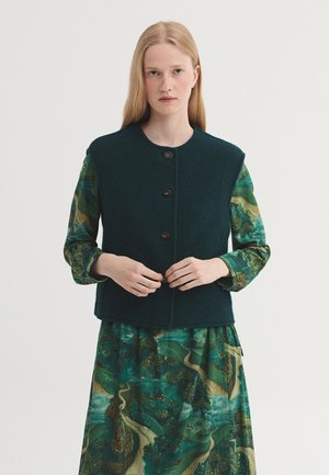 Young woman wearing a dark green buttoned sleeveless vest over a long-sleeve forest green dress with a landscape pattern.