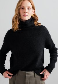 Black turtleneck sweater in soft, textured fabric with long sleeves, relaxed fit, and ribbed neckline worn over gray tailored trousers.