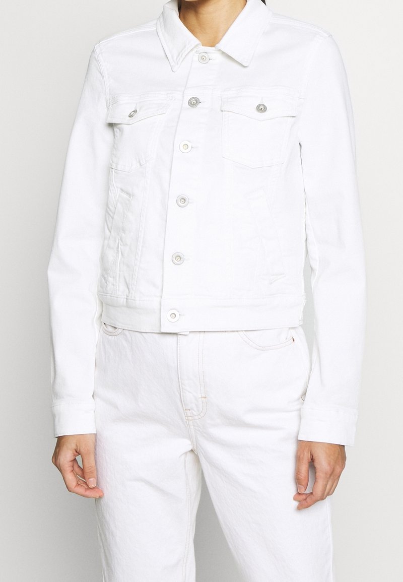 Chaqueta de denim blanca con un collar clásico, bolsillos frontales y herrajes plateados. Presenta un diseño ajustado y mangas largas. Textura suave.