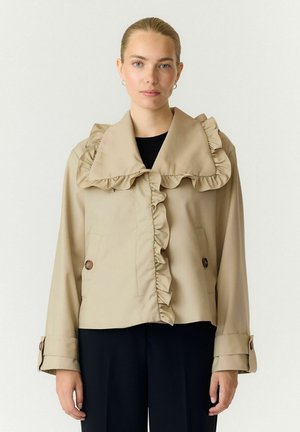 Donna con capelli tirati indietro che indossa una giacca beige con ampio colletto arricciato e davanti, abbinata a pantaloni neri, in piedi davanti a uno sfondo semplice.