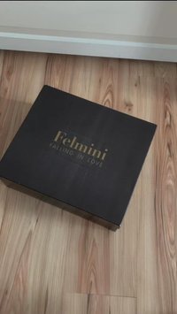 Černá krabička se zlatým textem "Felmini FALLING IN LOVE", hladkým povrchem, čtvercovým tvarem, umístěná na dřevěné podlaze.