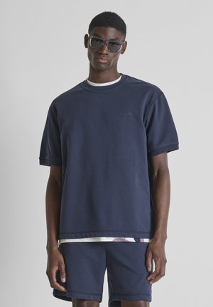 Uomo che indossa una T-shirt oversize blu navy, pantaloncini abbinati, occhiali da sole, in piedi davanti a uno sfondo grigio uniforme.