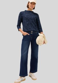 Maglia a maniche lunghe blu navy con motivo a strisce zebrate, abbinata a jeans a gamba larga blu scuro e sneakers beige, mentre tiene una borsa color crema.