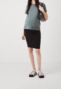 Mutterschafts-T-Shirt mit kurzen Ärmeln in gedämpftem Grün, kombiniert mit einem schwarzen, figurbetonten Rock. Die Schuhe bestehen aus schwarzen Sandalen mit weißen Riemen.