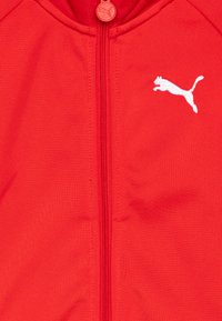 Veste zippée rouge en tissu lisse. Elle présente un logo blanc bien visible, un col rond et un bouton rouge assorti en haut.