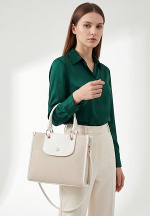 Femme en blouse verte et pantalon beige tenant un sac à main beige et blanc avec poignées supérieures et bandoulière.