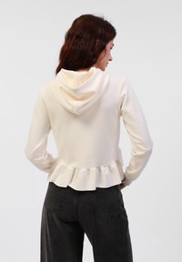 Felpa leggera color crema con orlo arricciato e cappuccio con cordino, realizzata in un tessuto morbido; abbinata a jeans neri a vita alta.