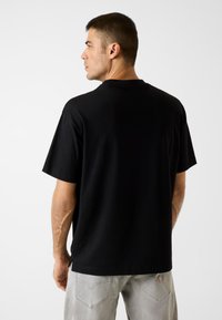 Schwarzes Baumwoll-T-Shirt mit kurzen Ärmeln und entspanntem Schnitt. Verfügt über einen geraden Saum und leichte Seitenschlitze. Die Rückansicht zeigt eine glatte Textur.