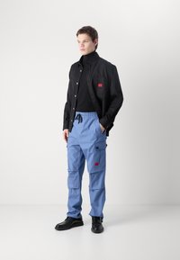 Camisa preta de botões com um rótulo vermelho, combinada com calças cargo azul claro com bolsos laterais e cintura ajustável por cordão. Sapatos pretos.