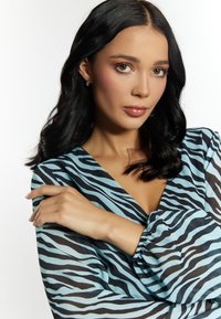 Blusa a strisce zebra blu e nera con scollatura a V profonda, maniche arricciate e un tessuto leggero e trasparente. Modella in posa con le braccia incrociate.