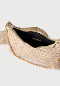 Sac bandoulière beige à motif avec une chaîne dorée, ouverture supérieure par fermeture éclair dévoilant un intérieur en tissu noir avec une poche intérieure zippée.