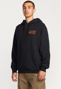 Sort hættetrøje med en orange "VOLCOM" logo og tekst på øverste venstre side. Inkluderer en frontlomme og ribbede manchetter.